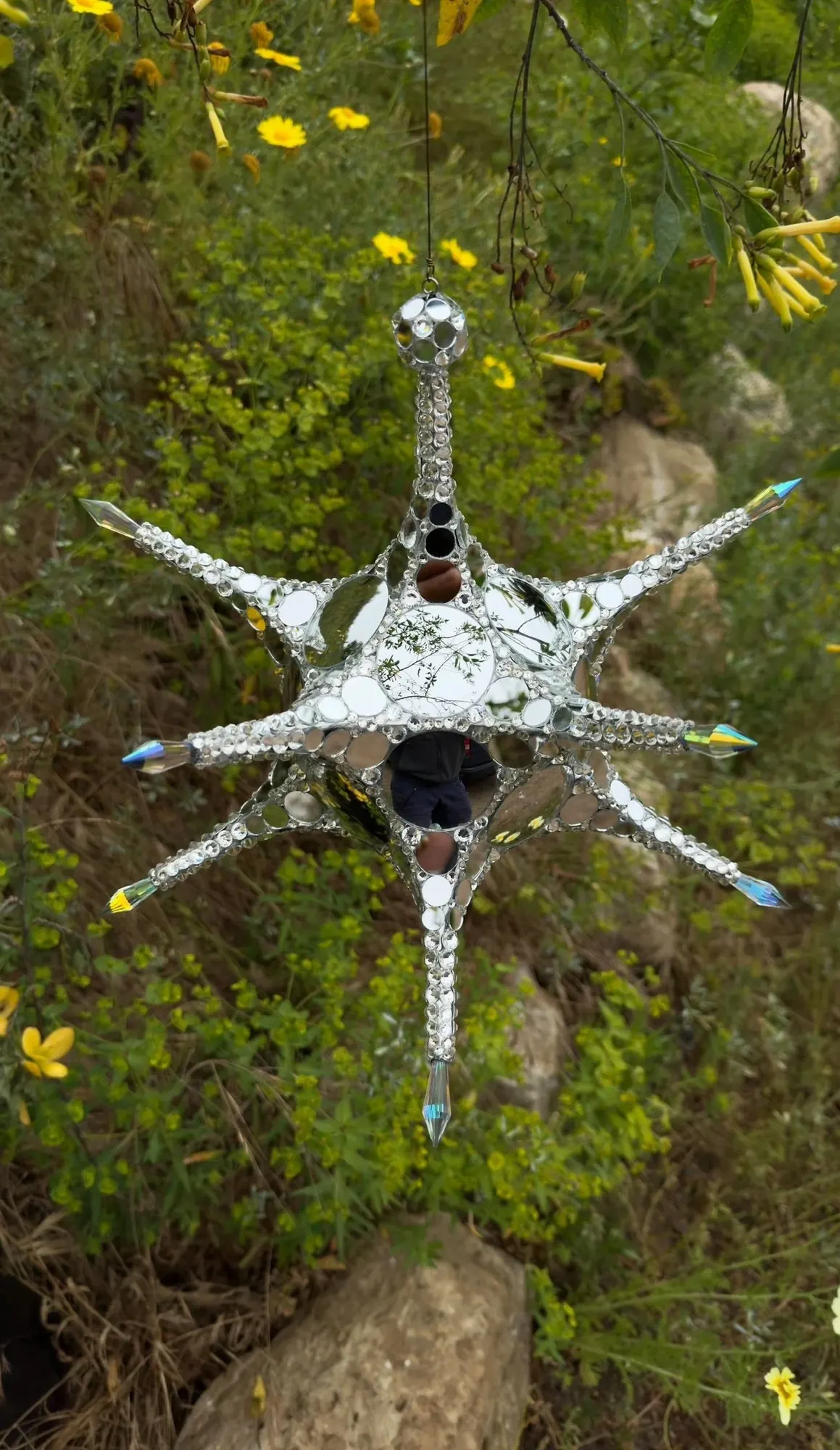 Metatron Reflections mirrored star sculpture hanging outdoors among yellow flowers