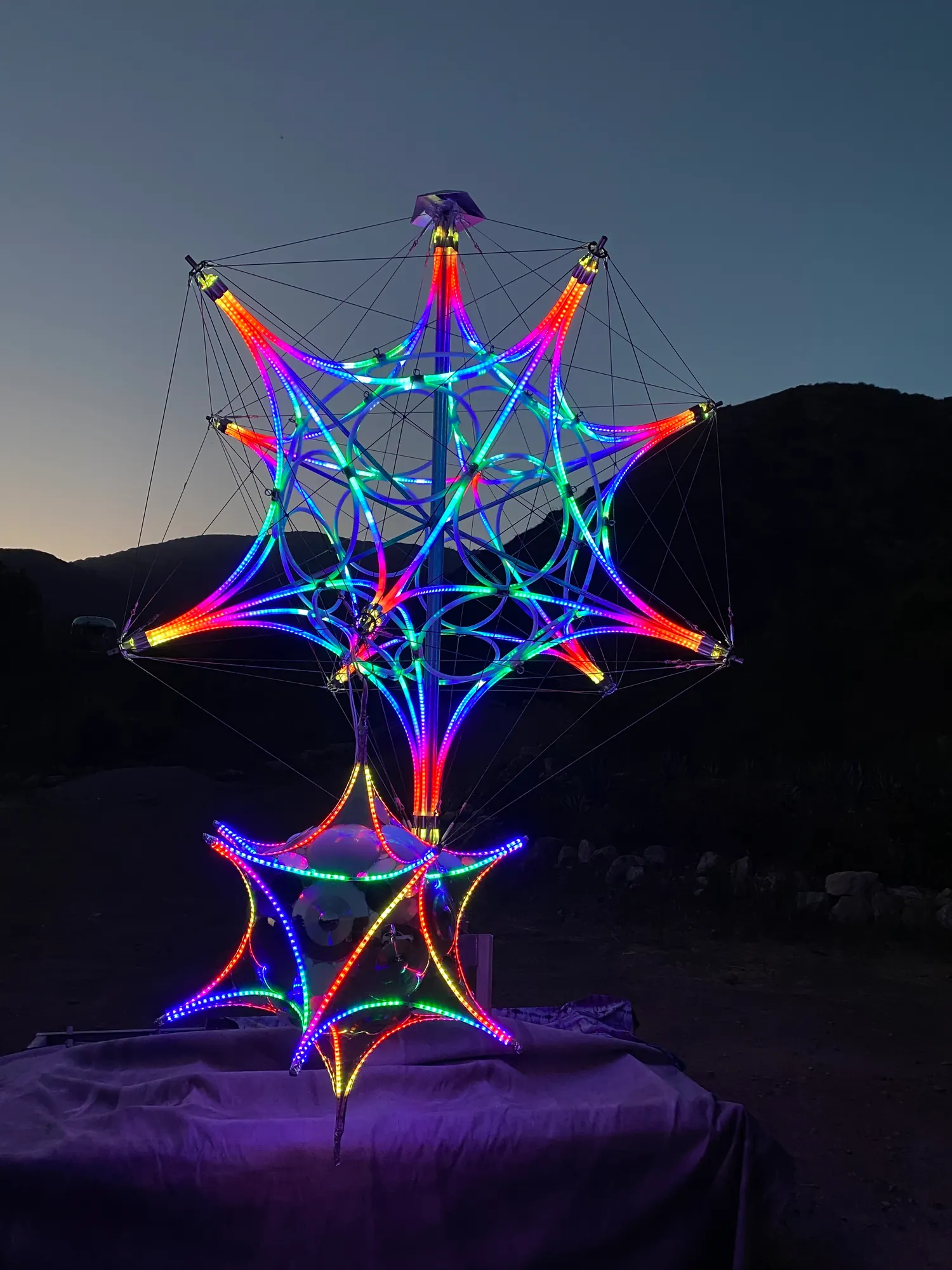 A large geometric star sculpture outlined in coloured light at night, standing on a low platform
