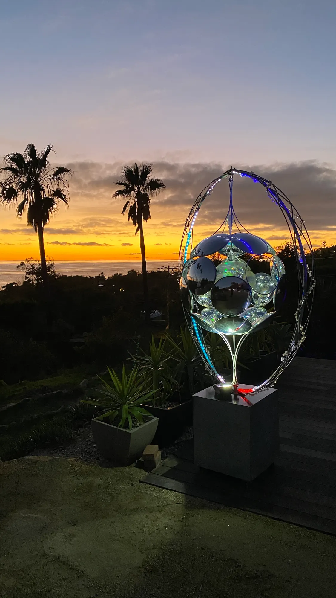 A mirrored sphere held in a tall metal ring on a stone plinth, lit against a sunset sky and palms