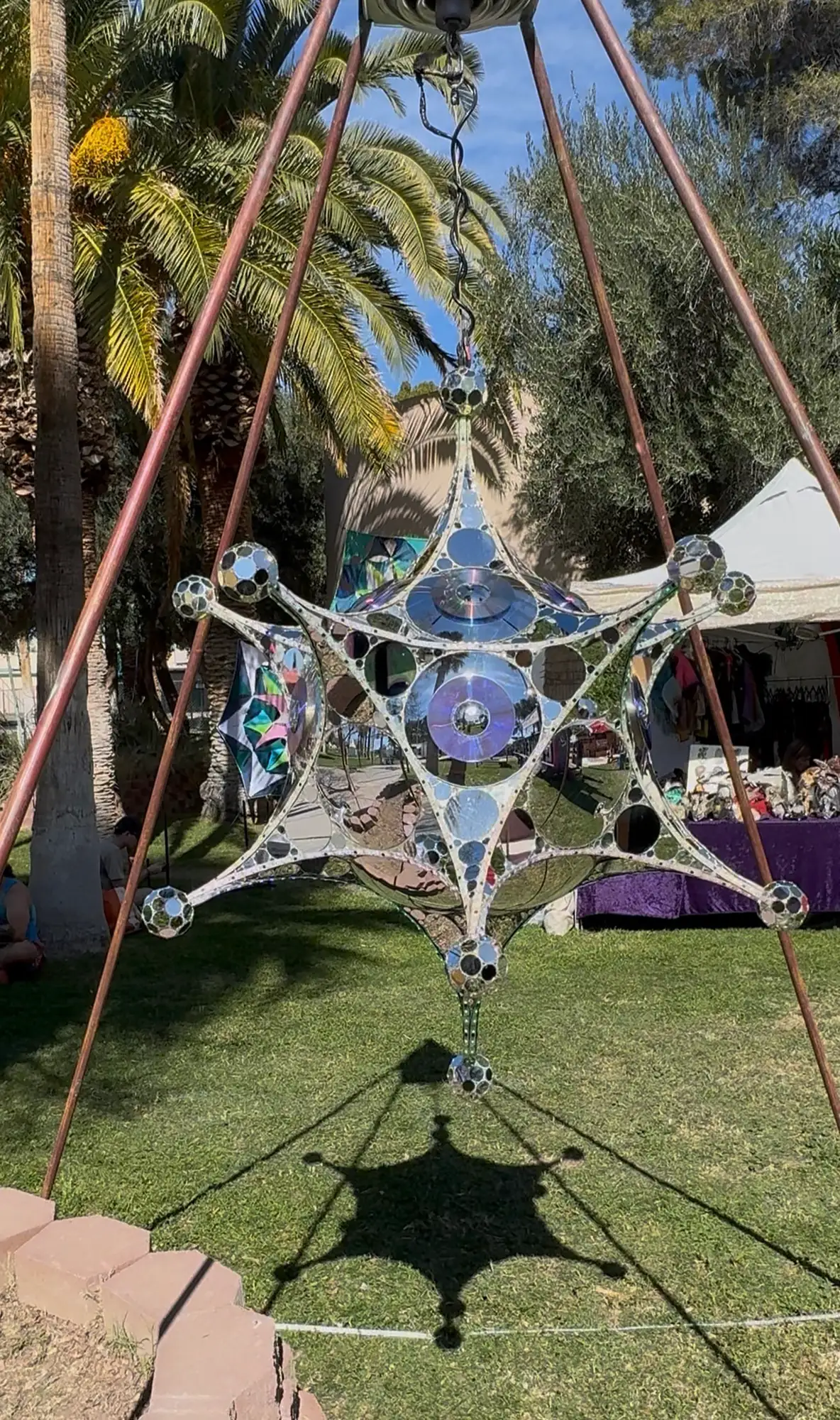 A mirrored star sculpture suspended from a copper tripod over a lawn, casting a star-shaped shadow