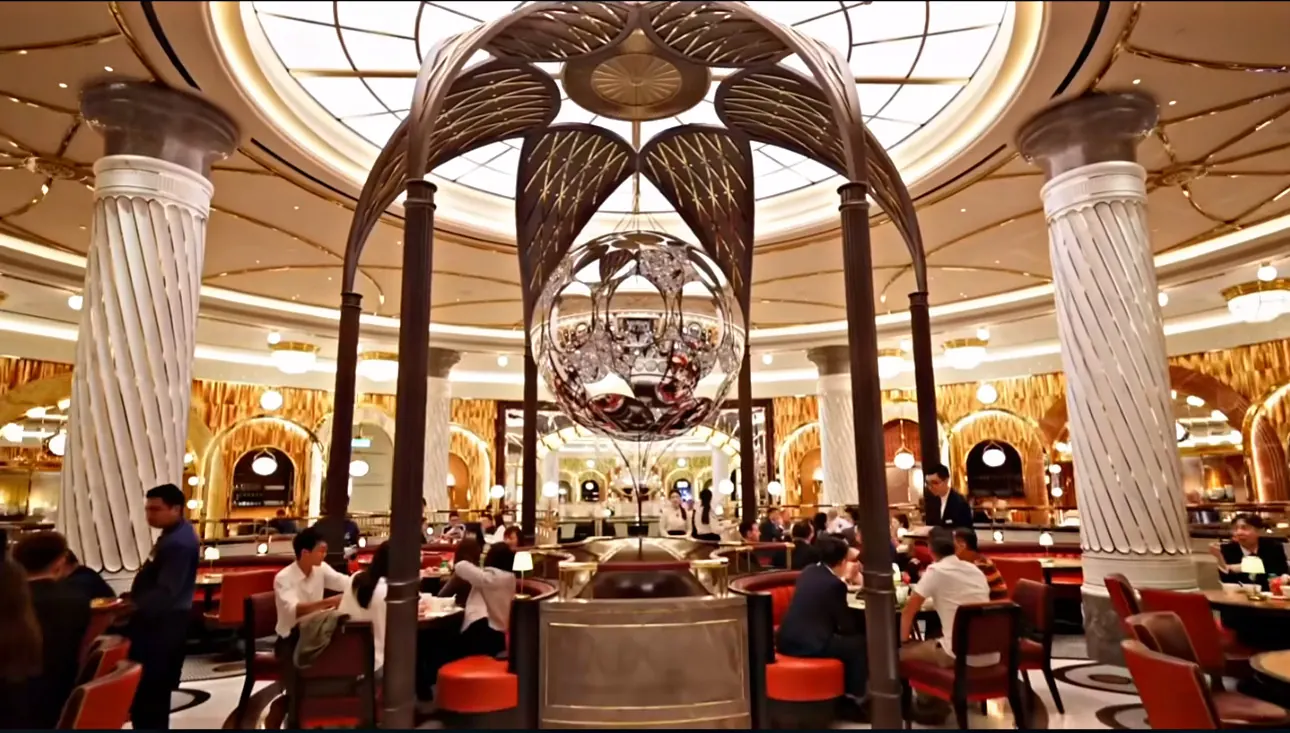 A mirrored sphere hanging under a bronze canopy at the centre of an ornate restaurant, with a glass dome above and diners at tables around it