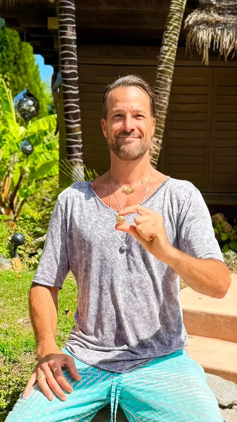 Nathan Skyhorse smiling in a sunlit garden before a dark wooden house, holding up a small gold sphere pendant on a chain around his neck, with mirrored spheres hanging among banana leaves at the left