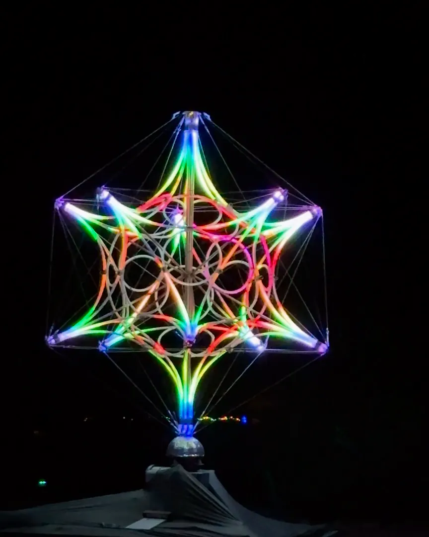 The All-One Sphyr, a large star of LED-tube rings on a draped platform, spins at night while its lights chase through colour.
