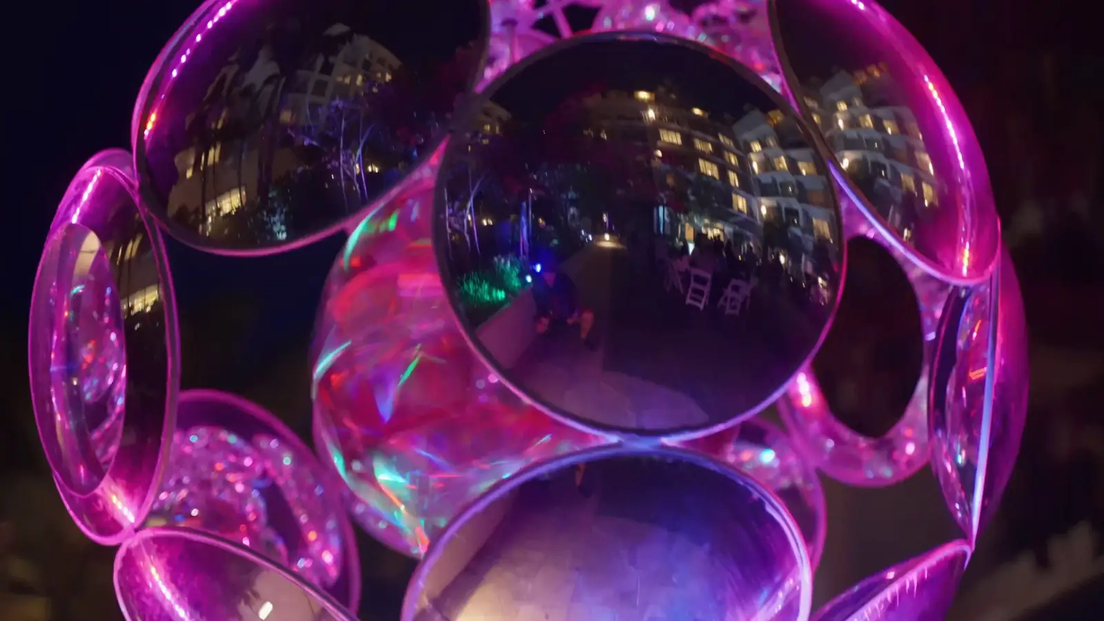 At night the camera tilts down through the BluRey AllSphyr, a sphere of clear convex lenses in an LED ring, as its light cycles from green and blue through purple and pink, the lit hotel reflected in the lenses.